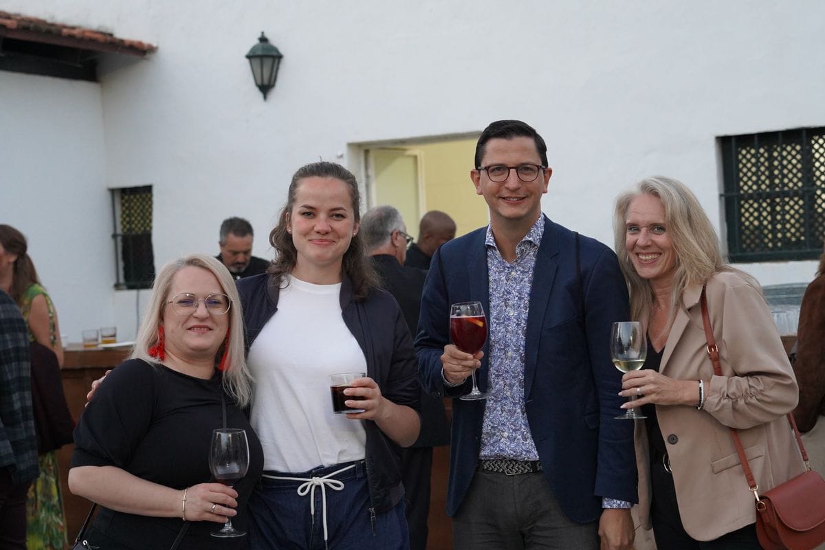 Vier Personen stehen im Freien vor einer weißen Wand und halten Weingläser in den Händen. Sie lächeln in die Kamera, während im Hintergrund weitere Gäste sind.