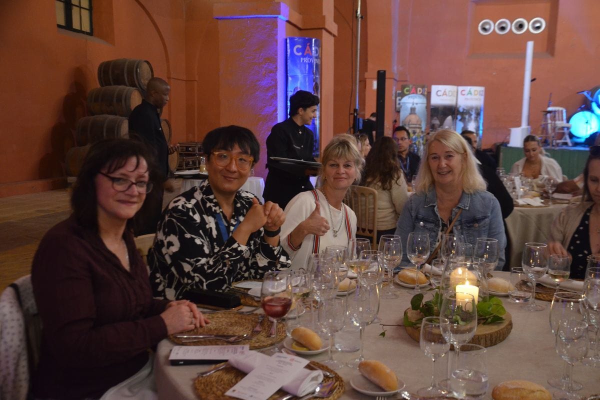Eine Gruppe von Menschen sitzt an einem festlich gedeckten runden Tisch in einem Raum mit orangefarbenen Wänden und Weinfässern im Hintergrund.