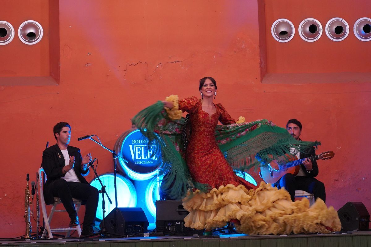 Eine Flamenco-Tänzerin in einem rot-gelben Kleid mit grünem Tuch tanzt auf einer Bühne vor zwei Musikern, die Gitarre spielen und in die Hände klatschen.