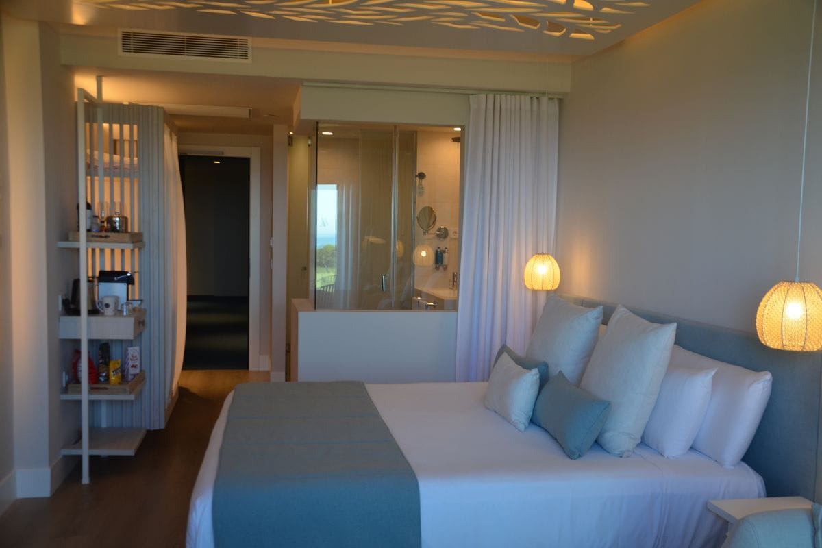 Ein modernes Hotelzimmer mit einem großen Doppelbett, einer offenen Glasduschkabine, einem Regal mit Minibar und zwei hängenden Lampen an der Decke.