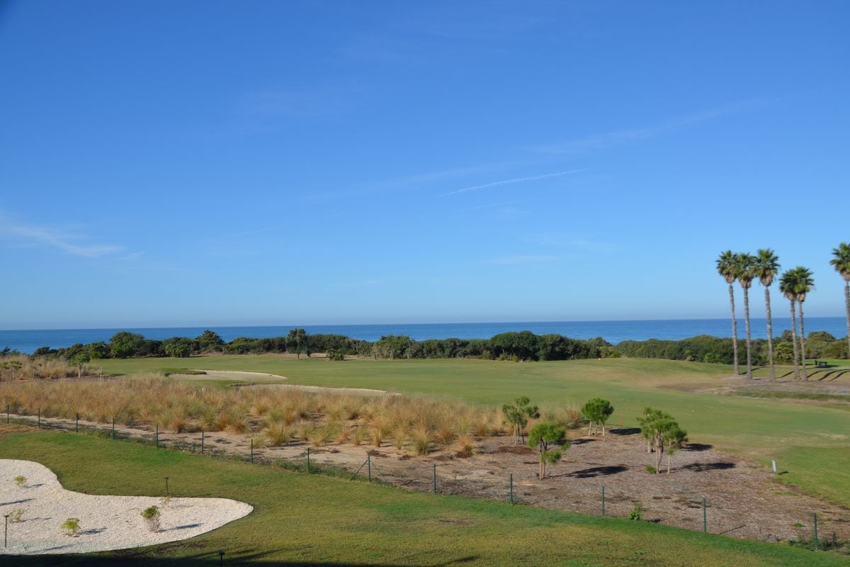 Blick auf einen Golfplatz mit Grünflächen, Sandbunkern und Palmen vor einem tiefblauen Meer unter einem wolkenlosen, hellblauen Himmel an einem Sonnentag.