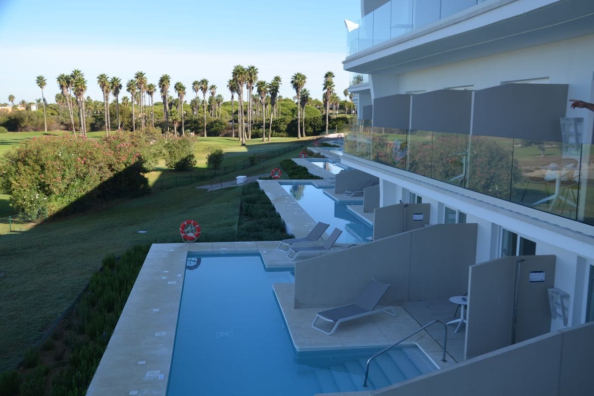 Blick von einem Hotelbalkon auf eine Reihe von privaten Terrassen mit kleinen Pools und Liegestühlen vor einer weitläufigen Grünanlage mit Palmen.