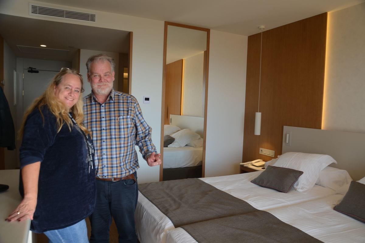 Eine blonde Frau und ein Mann mit Bart in einem modern eingerichteten Hotelzimmer, das mit einem großen Doppelbett und einer Holzvertäfelung ausgestattet ist.