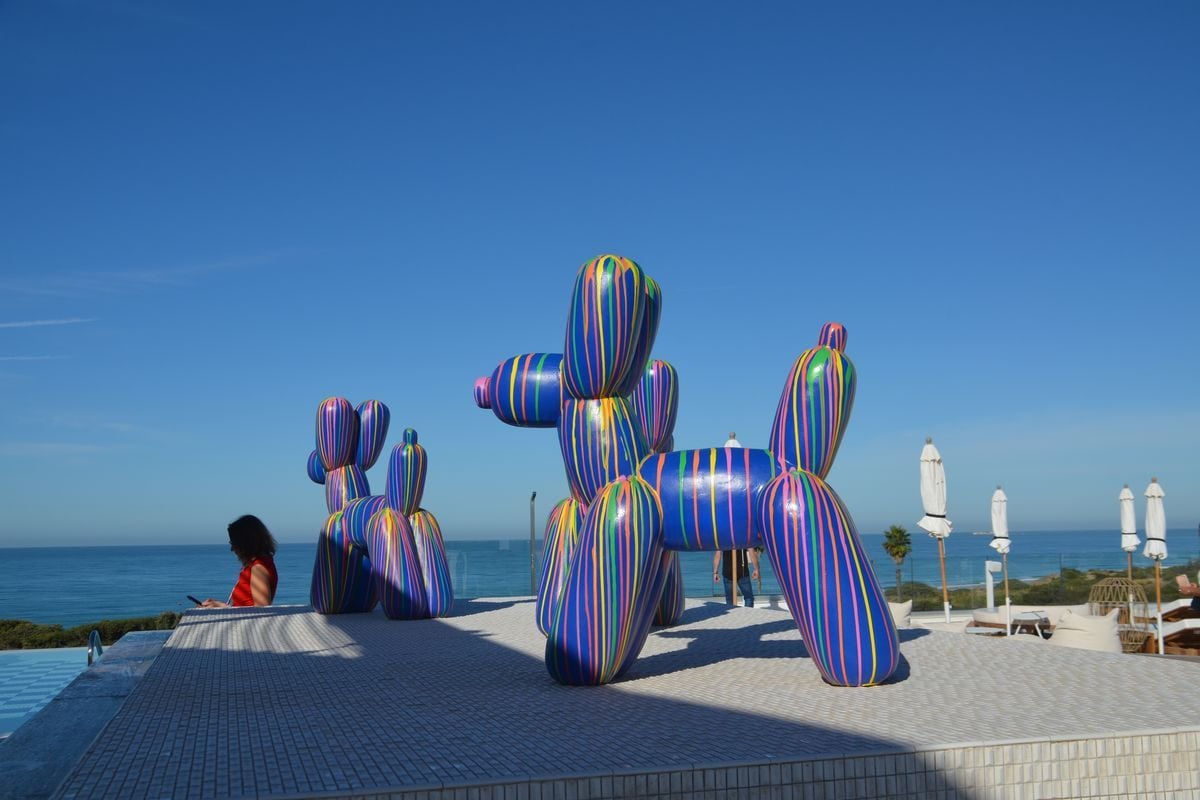 Zwei große, bunt gestreifte Skulpturen in Form von Ballonhunden stehen auf einer gefliesten Terrasse vor dem Hintergrund eines blauen Meeres und Himmels.