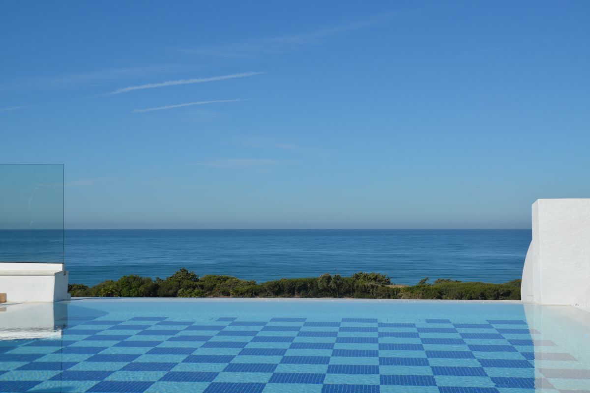 Ein Infinity-Pool mit blau-weißem Schachbrettmuster im Vordergrund, dahinter eine grüne Küstenlandschaft und der weite, blaue Ozean unter klarem Himmel.