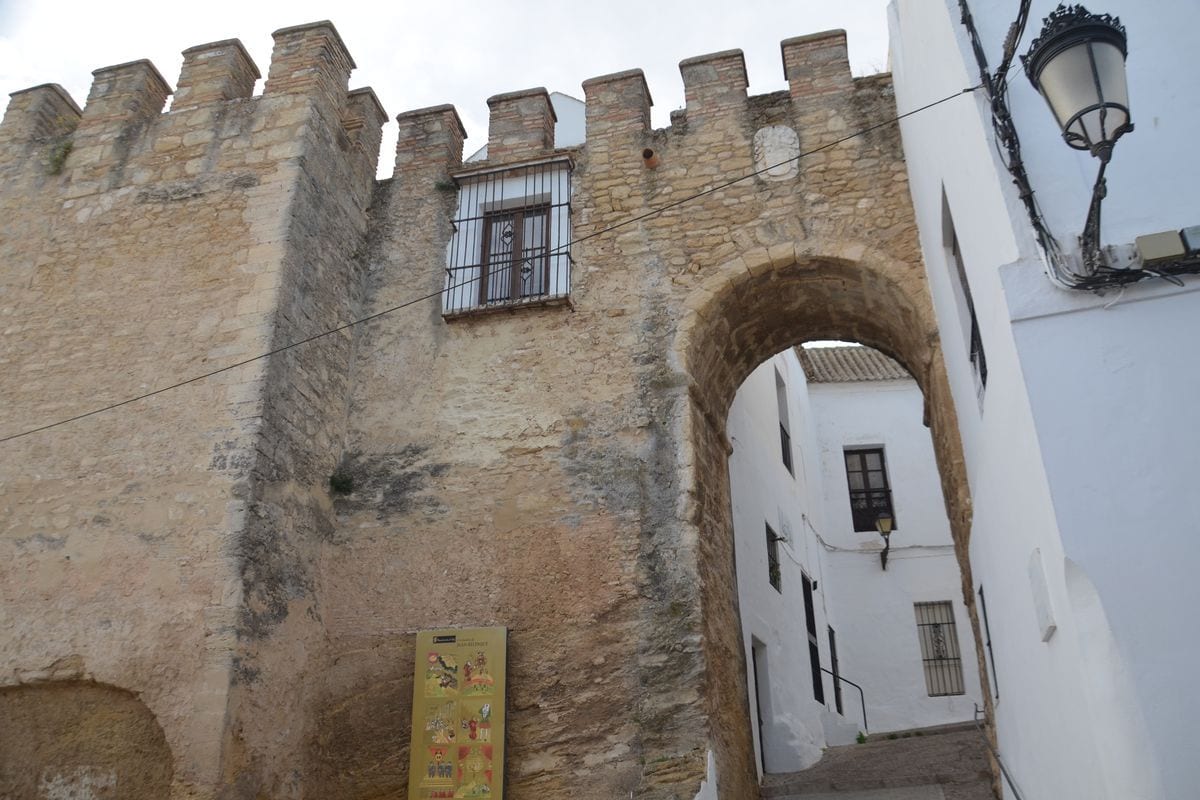 Ein historisches Stadttor aus Naturstein mit Zinnen und einem Torbogen, das zu einer weiß getünchten Gasse mit einer Straßenlaterne und Fenstern führt.
