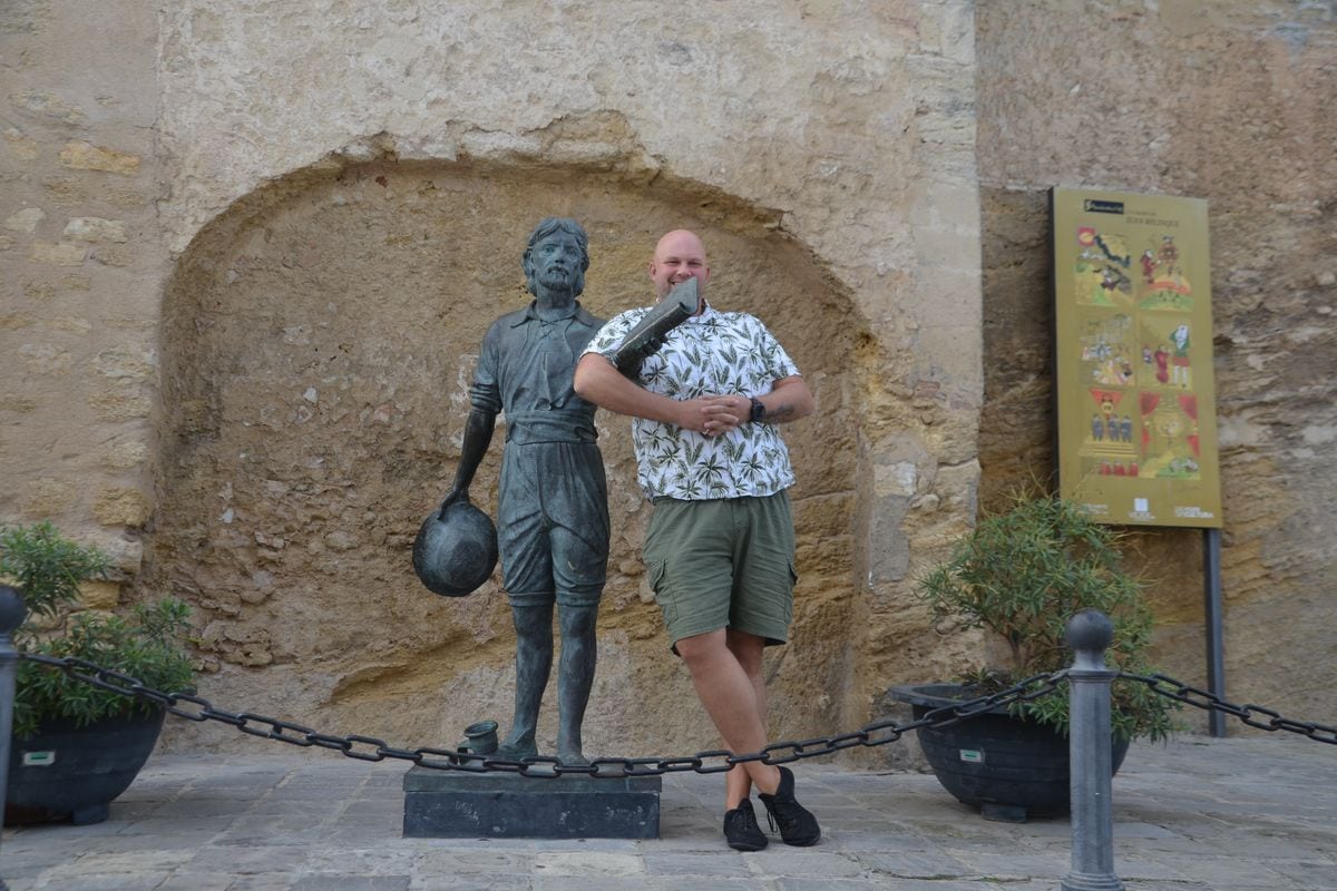 Ein Mann mit Glatze und gemustertem Hemd lehnt lächelnd an einer bronzenen Statue vor einer historischen Steinmauer mit einer Informationstafel daneben.