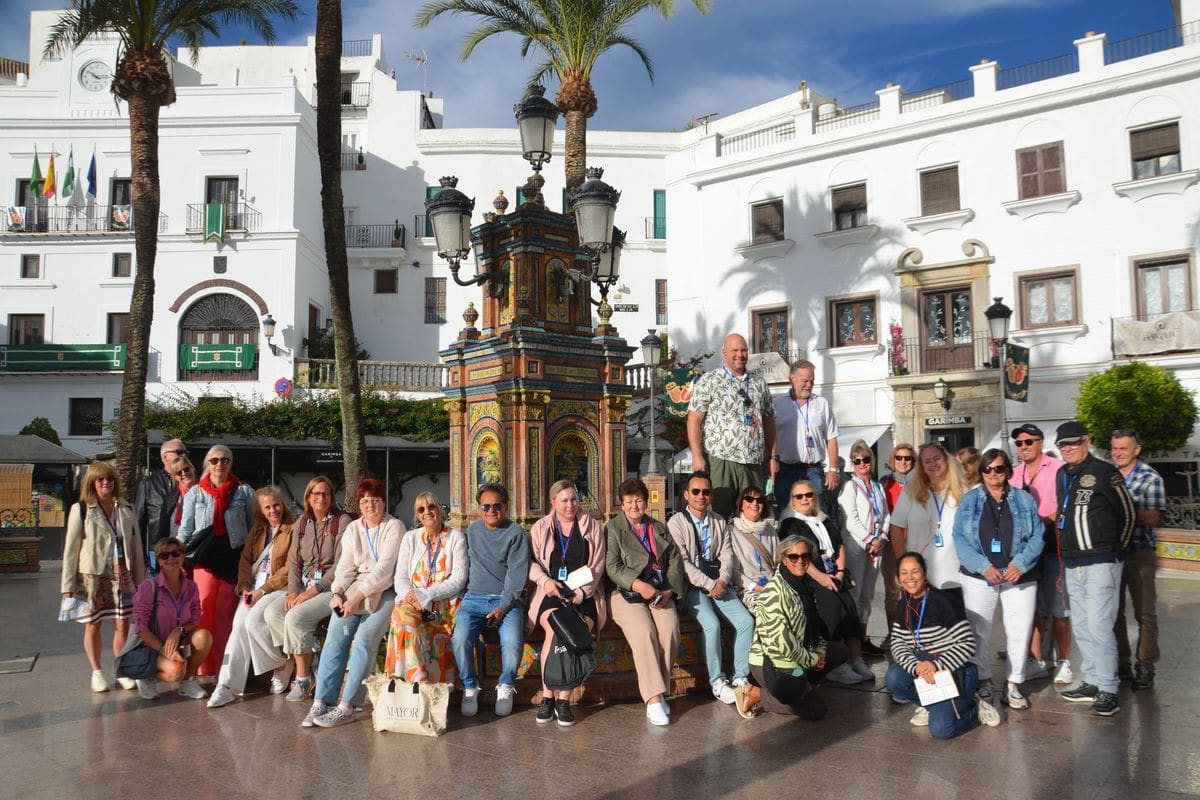 Eine große Gruppe von Menschen posiert für ein Foto auf einem sonnigen Platz vor einem verzierten Brunnen und weißen Gebäuden in einer spanischen Stadt.