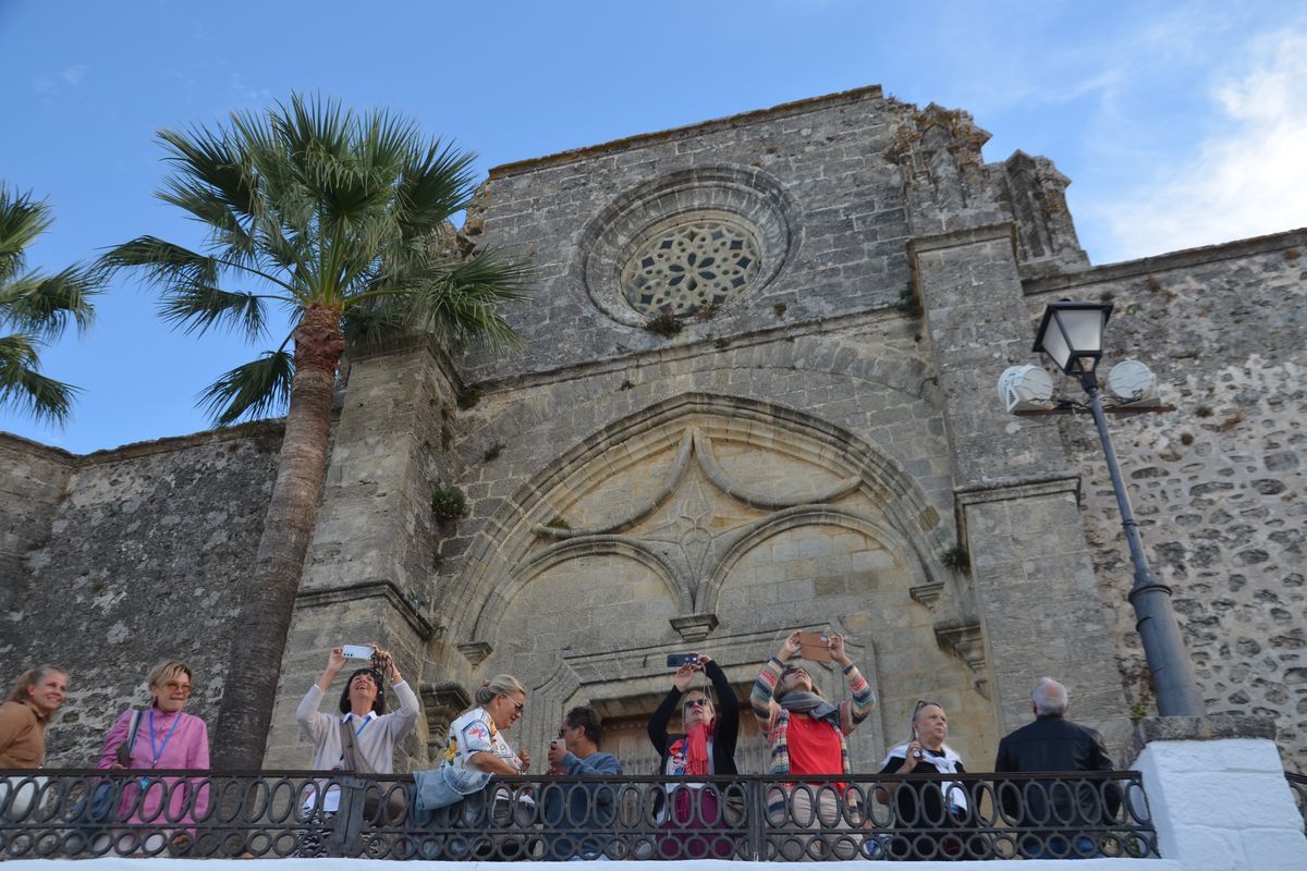 Eine Gruppe von Menschen steht vor einer historischen Steinfassade mit Rosettenfenster und fotografiert diese mit ihren Smartphones unter einer Palme.