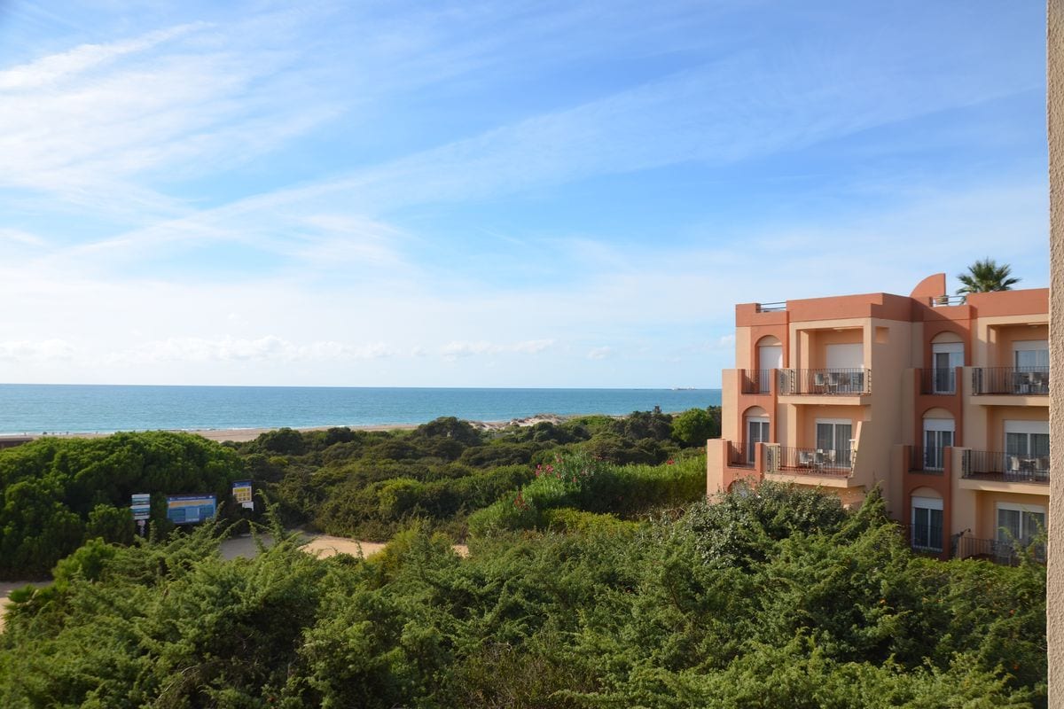 Blick von einem erhöhten Standpunkt auf ein mehrstöckiges Hotelgebäude, das von dichter Vegetation umgeben ist und direkt an einen Sandstrand grenzt.