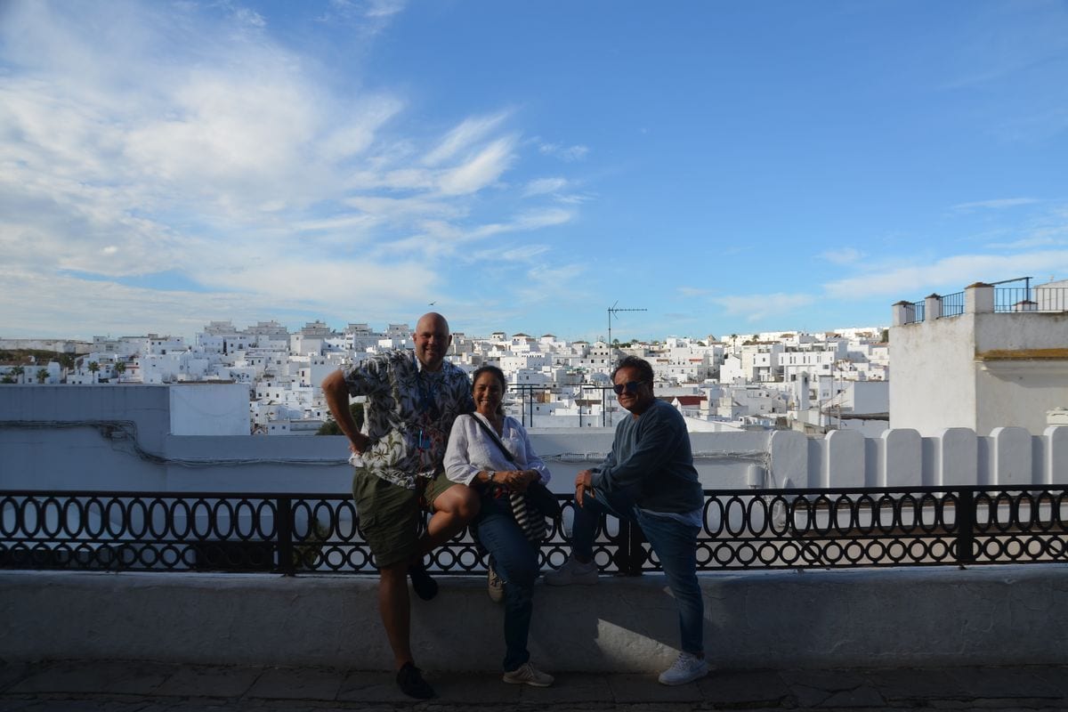 Drei Personen stehen vor einem Geländer mit Blick auf eine weiße Stadt in Spanien, im Hintergrund erstreckt sich eine Hügellandschaft unter blauem Himmel.
