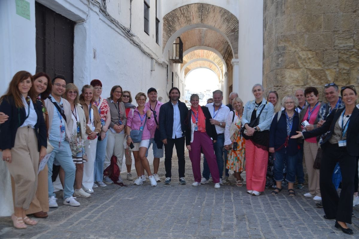 Eine große Gruppe von Menschen steht in einer schmalen, gepflasterten Gasse zwischen weißen Gebäuden und einem gemauerten Torbogen für ein Foto zusammen.