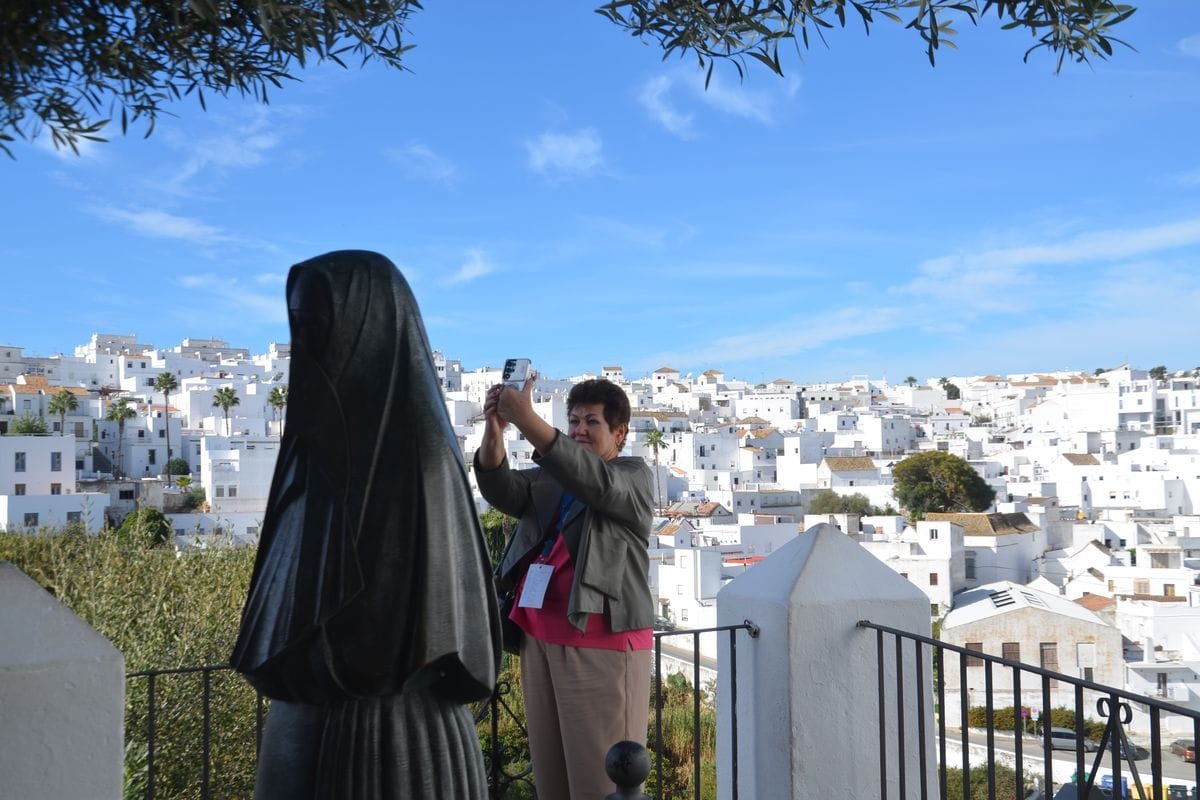 Eine Frau fotografiert mit ihrem Smartphone eine dunkle Bronzestatue vor der Kulisse einer weißen Stadt in Spanien unter einem strahlend blauen Himmel.