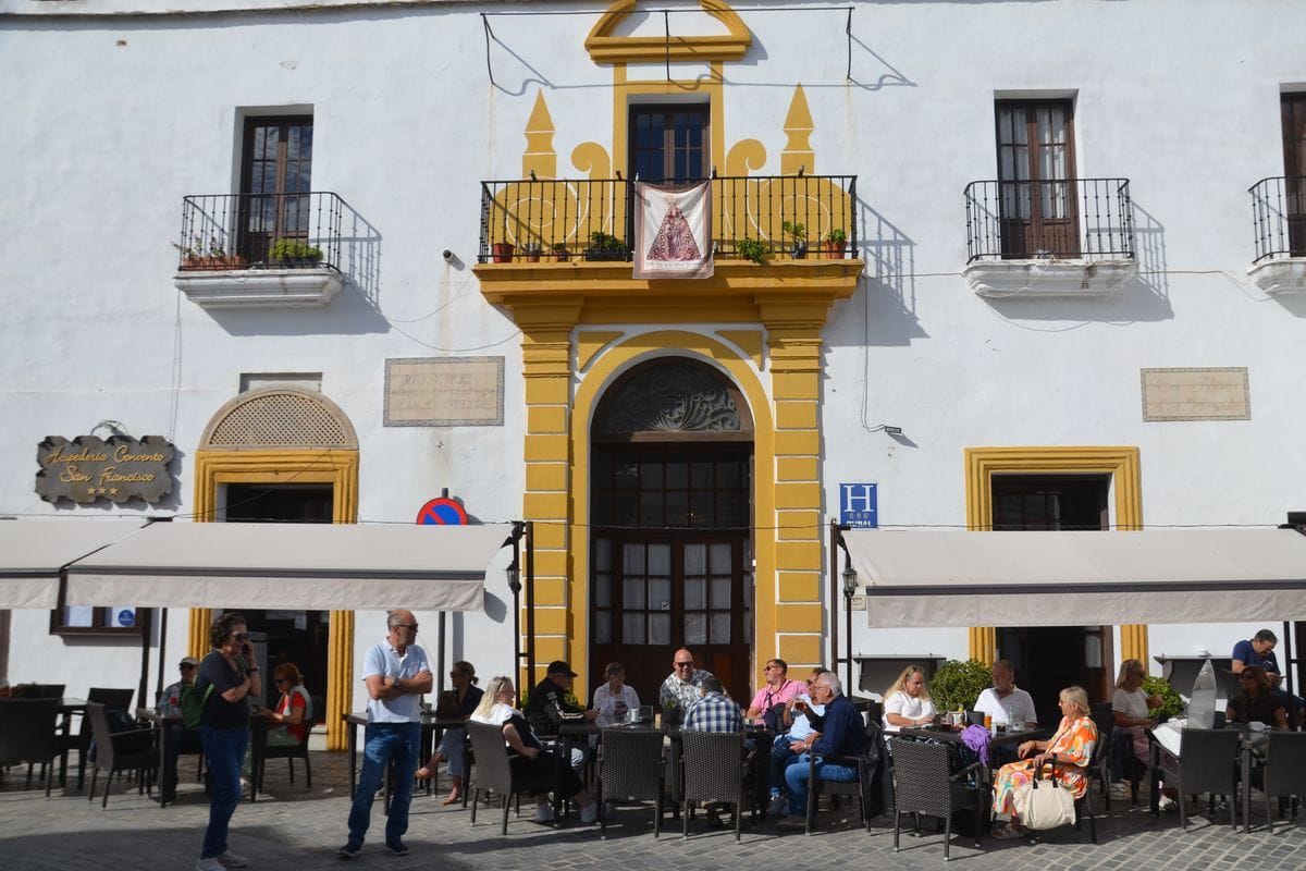 Die weiße Fassade der Hospederia Convento San Francisco mit gelben Verzierungen, Balkonen und Gästen an Tischen auf dem sonnigen Vorplatz in Spanien.