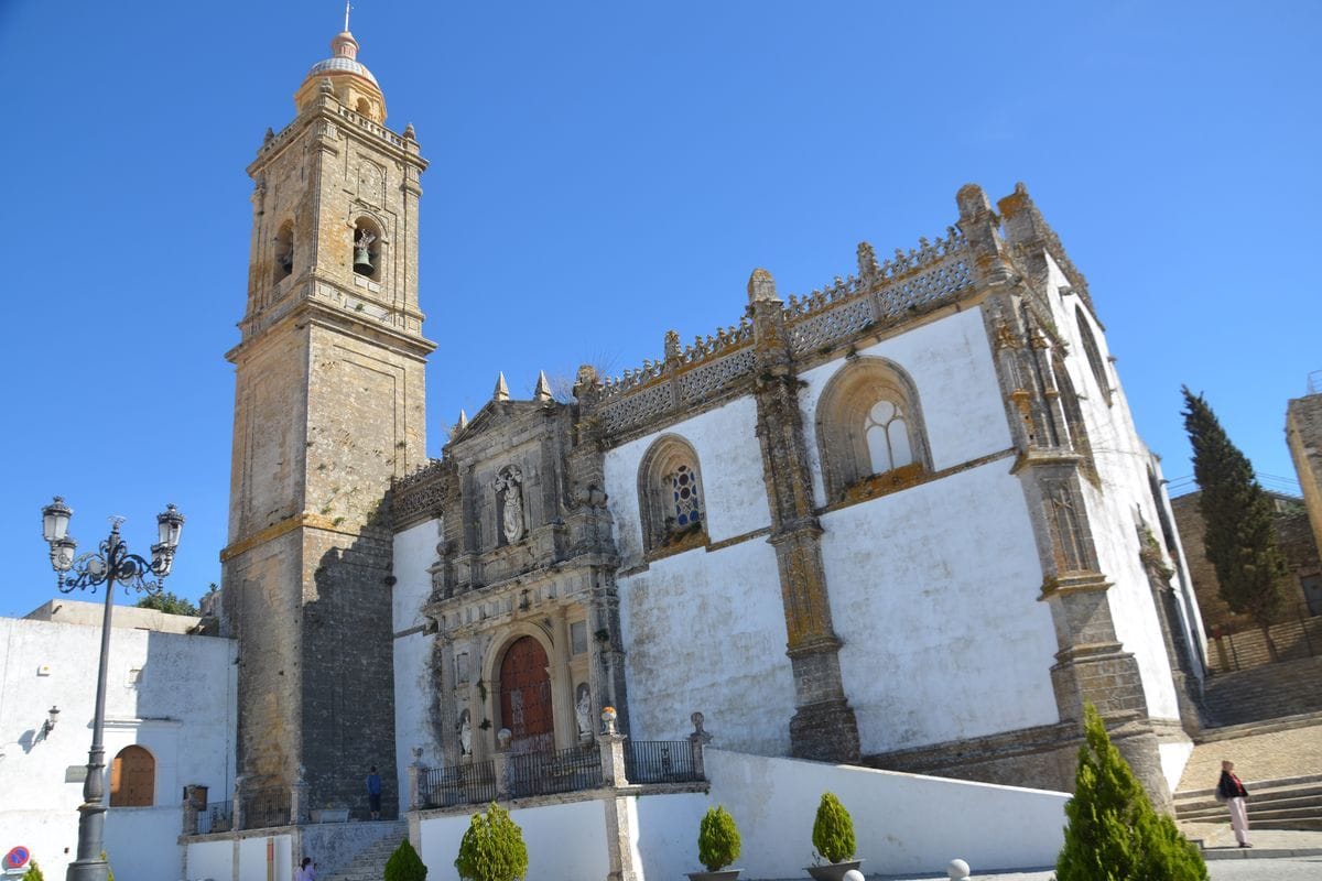 Eine historische Kirche mit hohem Glockenturm und weißer Fassade unter blauem Himmel, davor ein schmiedeeiserner Laternenmast und kleine grüne Ziersträucher.