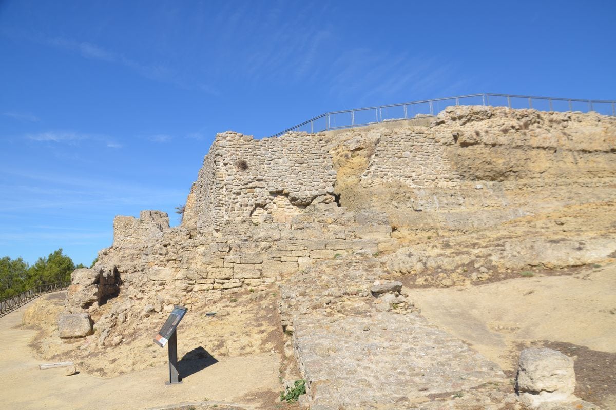 Historische Steinruinen auf einer Anhöhe unter blauem Himmel, mit einem Metallgeländer an der Oberkante und einer Informationstafel im Vordergrund links.