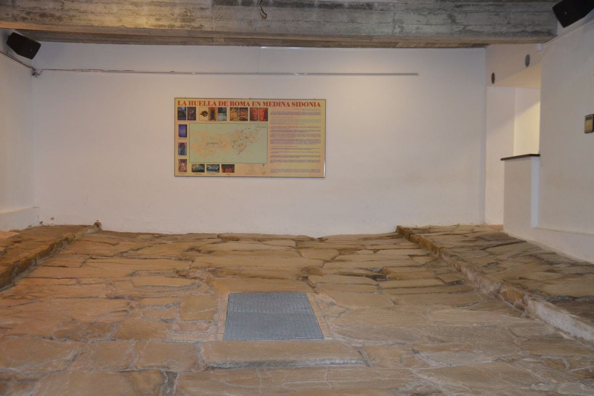 Ein Innenraum mit einem historischen Steinboden aus großen Platten und einer Informationstafel an der weißen Wand, die Medina Sidonia thematisiert.