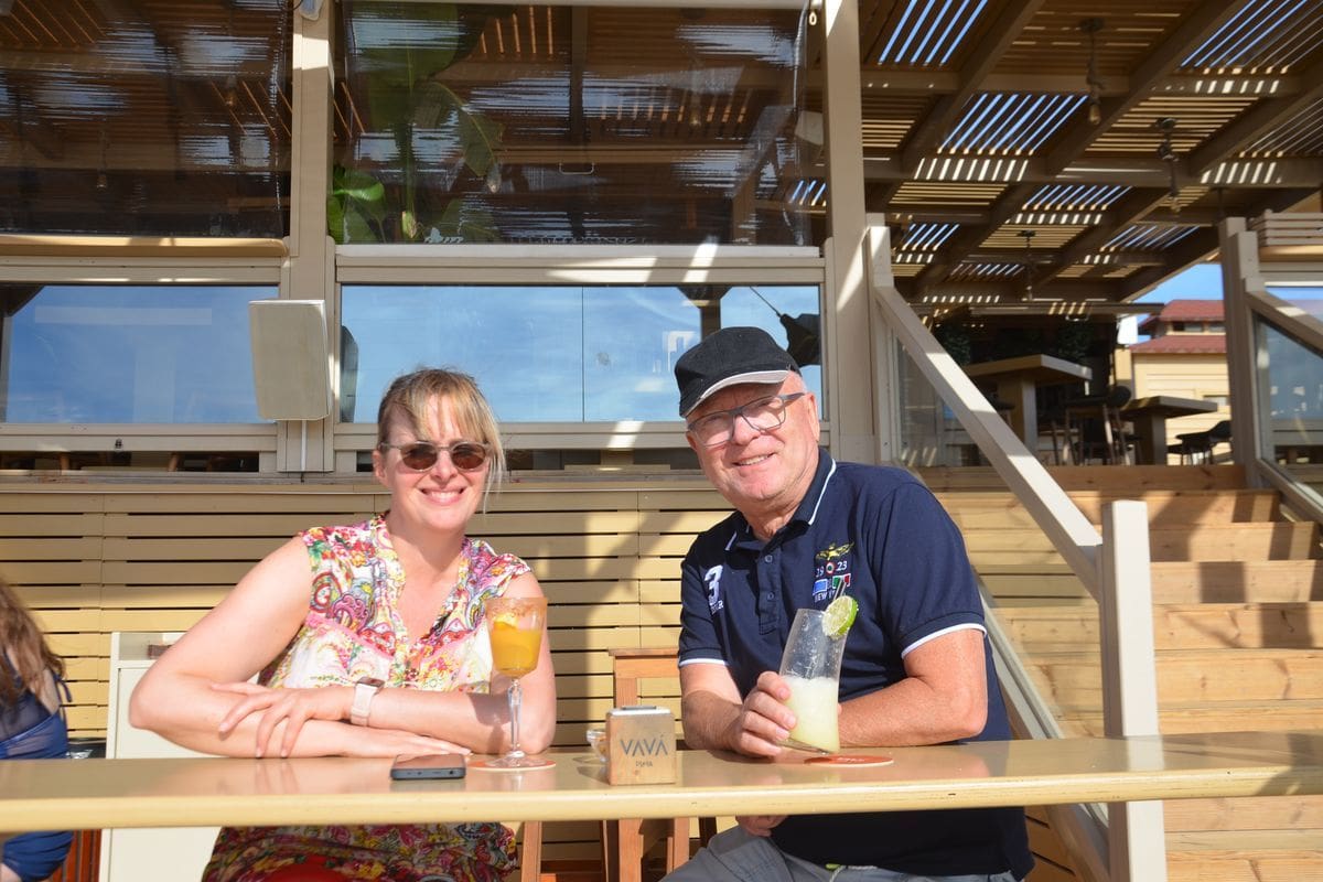 Eine Frau und ein Mann sitzen an einem Tisch auf einer Holzterrasse im Freien, halten jeweils ein Glas mit einem Getränk in der Hand und lächeln in Kamera.