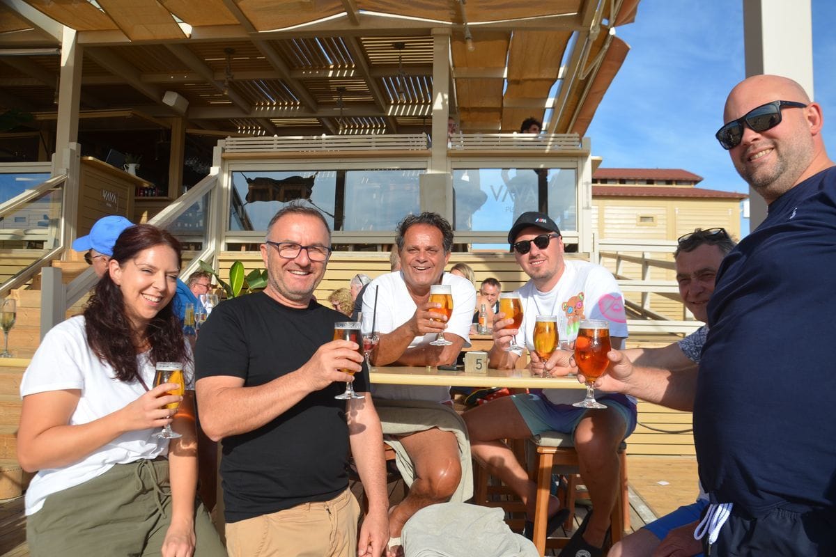 Eine Gruppe von sechs Personen sitzt an einem Tisch auf einer sonnigen Holzterrasse und hält jeweils ein Glas Bier in der Hand, während sie in die Kamera lächeln.