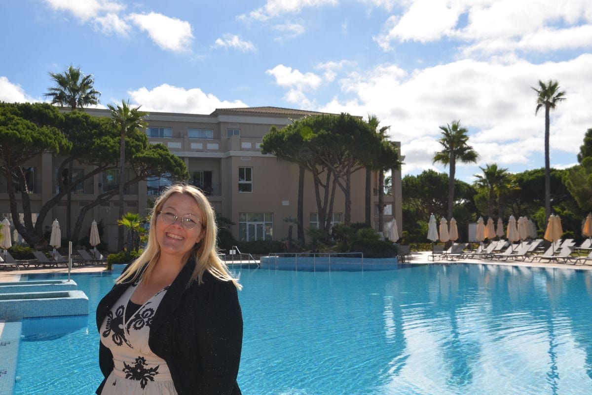 Eine lächelnde Frau mit Brille steht im Vordergrund vor einem großen Hotelpool, umgeben von Palmen, Liegestühlen und einem hellen Hotelgebäude im Hintergrund.