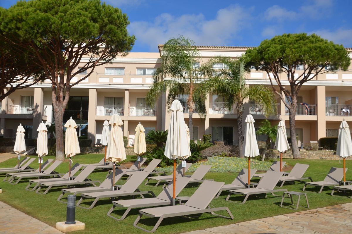 Eine Außenansicht eines Hotels mit beige gestrichener Fassade, umgeben von Pinien und Palmen, mit zahlreichen Sonnenliegen und geschlossenen Sonnenschirmen.