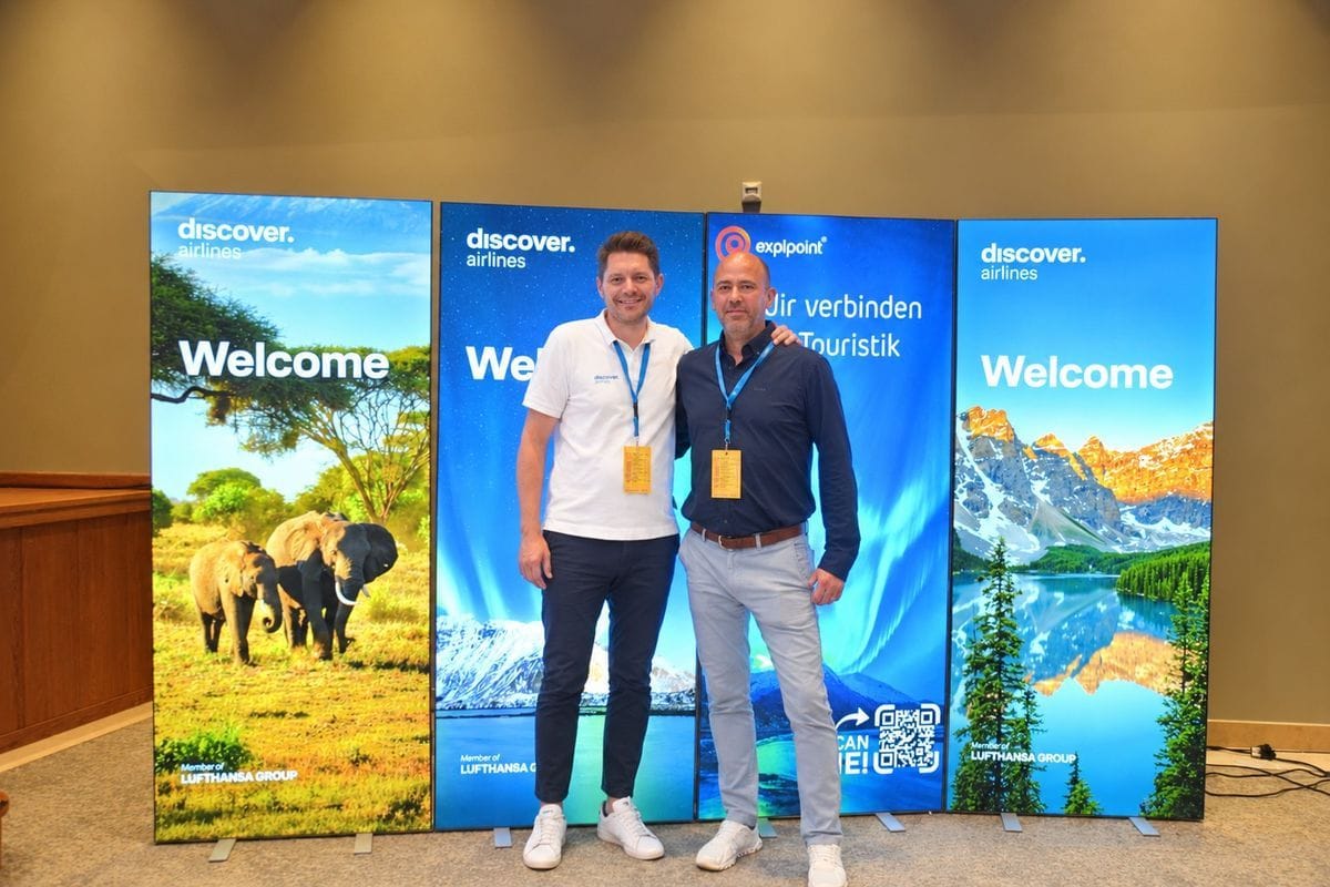 Zwei Männer stehen nebeneinander vor drei großen, beleuchteten Werbedisplays mit Naturmotiven und Logos von discover airlines und explpoint in einem Raum.