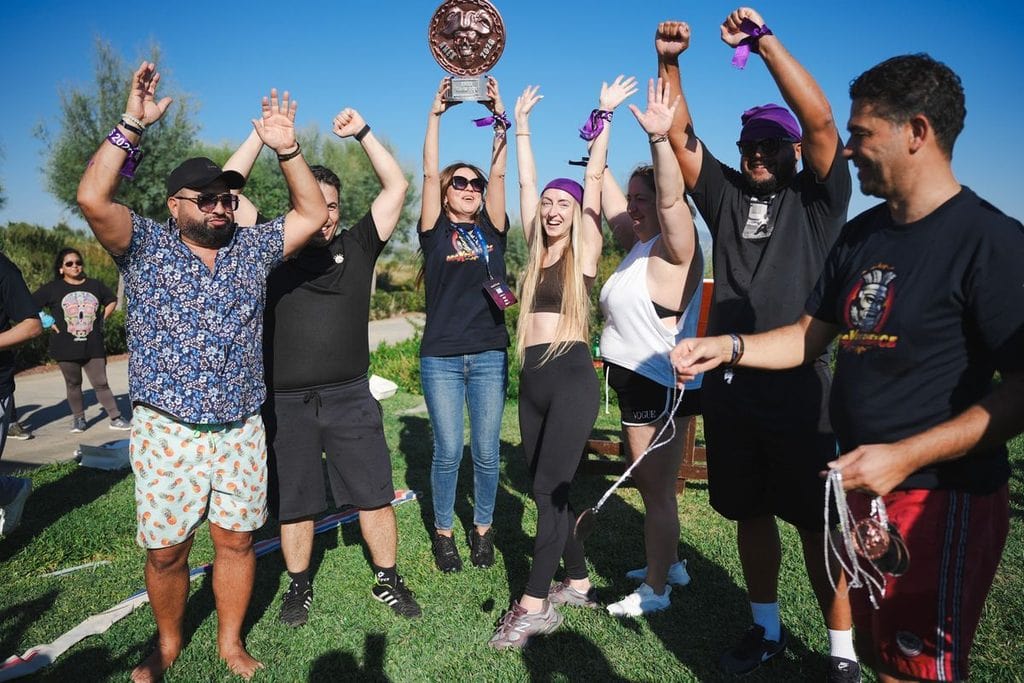 Eine Gruppe von Menschen steht im Freien auf einer Wiese, jubelt mit erhobenen Armen und hält einen goldenen Pokal sowie Medaillen in die Kamera.