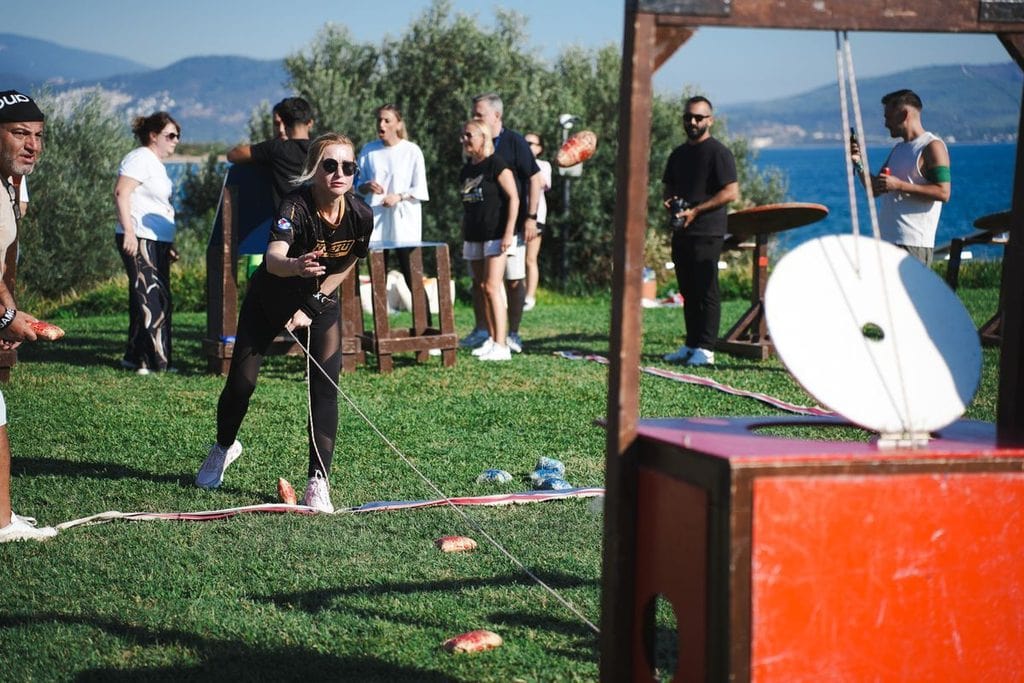 Eine Frau in Sportkleidung zieht an einem Seil, das mit einer Konstruktion verbunden ist, während eine Gruppe von Menschen im Hintergrund zuschaut.