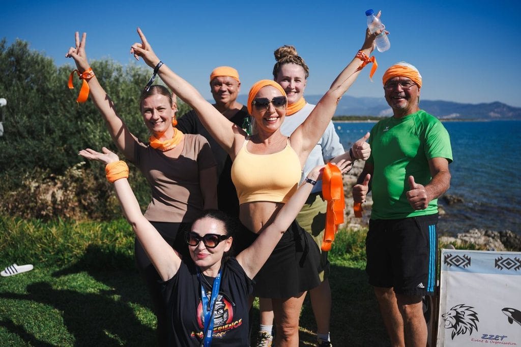 Eine Gruppe von sechs Personen steht im Freien vor dem Meer, trägt orangefarbene Stirnbänder und Armbänder und posiert fröhlich mit erhobenen Armen.