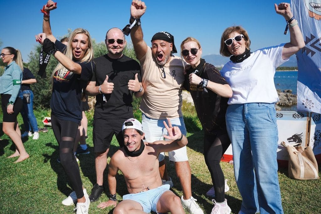 Eine Gruppe von sechs Personen posiert im Freien auf einer Wiese vor einer Küstenlandschaft, einige heben jubelnd die Arme und zeigen Daumen nach oben.