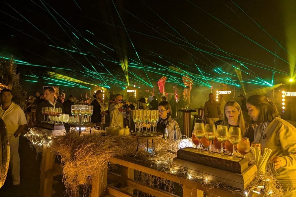Eine abendliche Veranstaltung im Freien mit einer Bar aus Holz, die mit Stroh und Lichterketten dekoriert ist, unter einem grünen Laserlicht-Himmel.