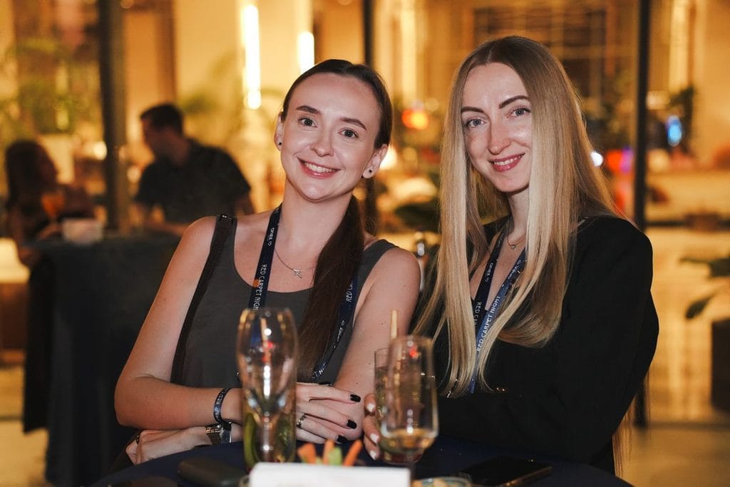 Zwei junge Frauen mit Lächeln und Umhängebändern um den Hals sitzen an einem Stehtisch mit Getränken in einer hell erleuchteten Hotel-Lobby am Abend.