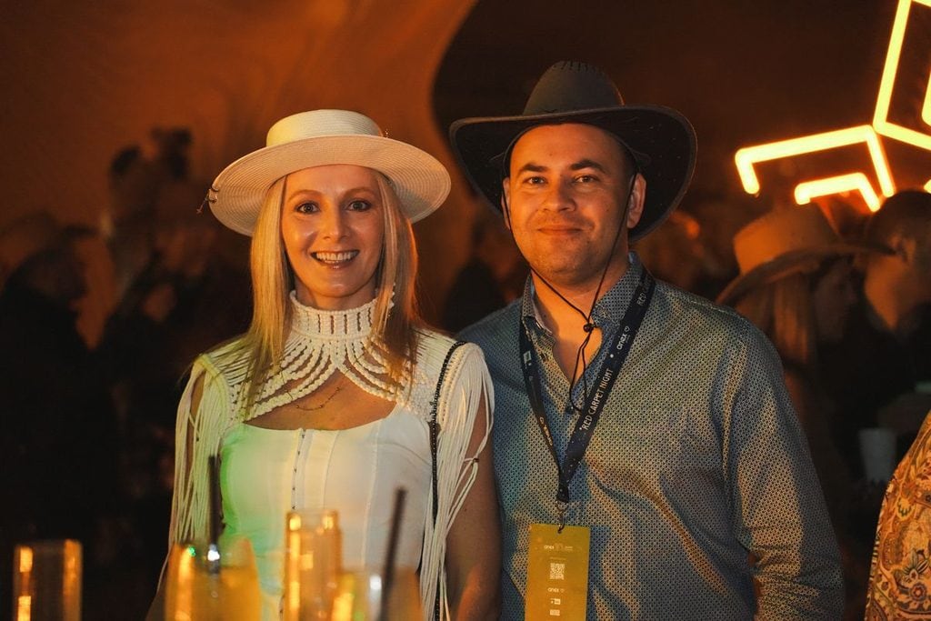 Eine Frau mit hellem Hut und ein Mann mit Cowboyhut stehen nebeneinander in einer abendlichen Umgebung mit warmem Licht und leuchtenden Neon-Elementen.