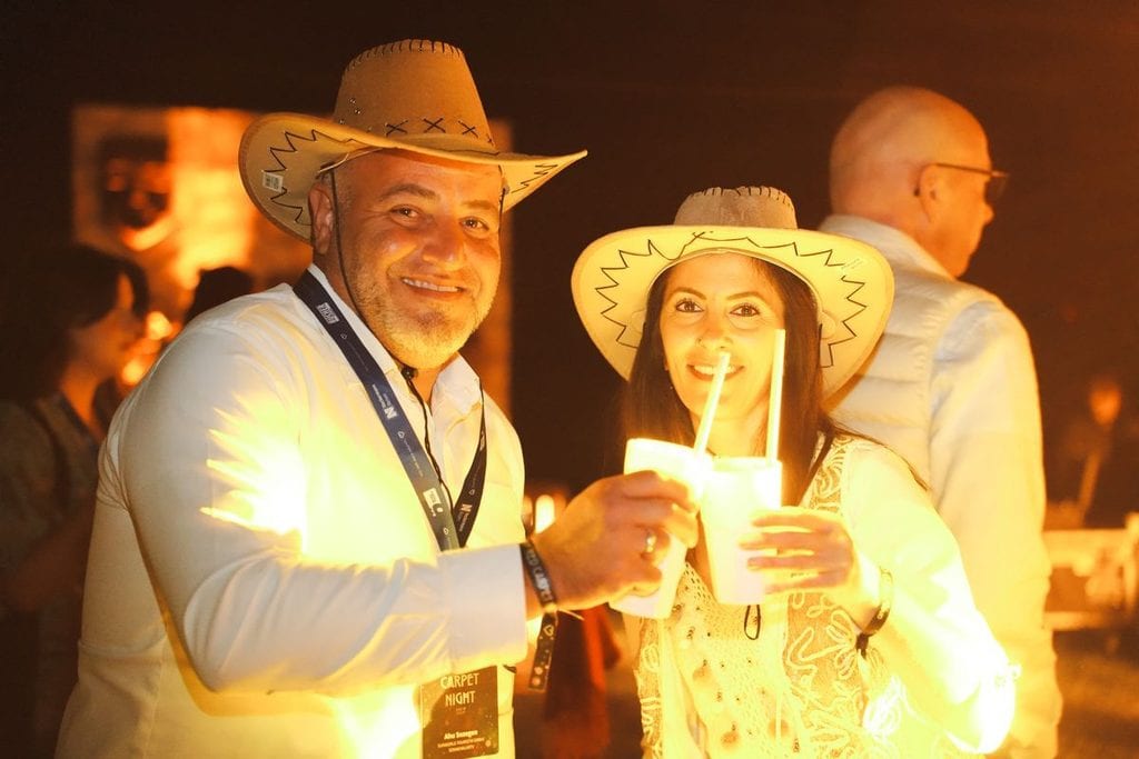 Ein Mann und eine Frau tragen Cowboyhüte und stoßen bei einer abendlichen Veranstaltung mit leuchtenden Getränkebechern in die Kamera lächelnd an.