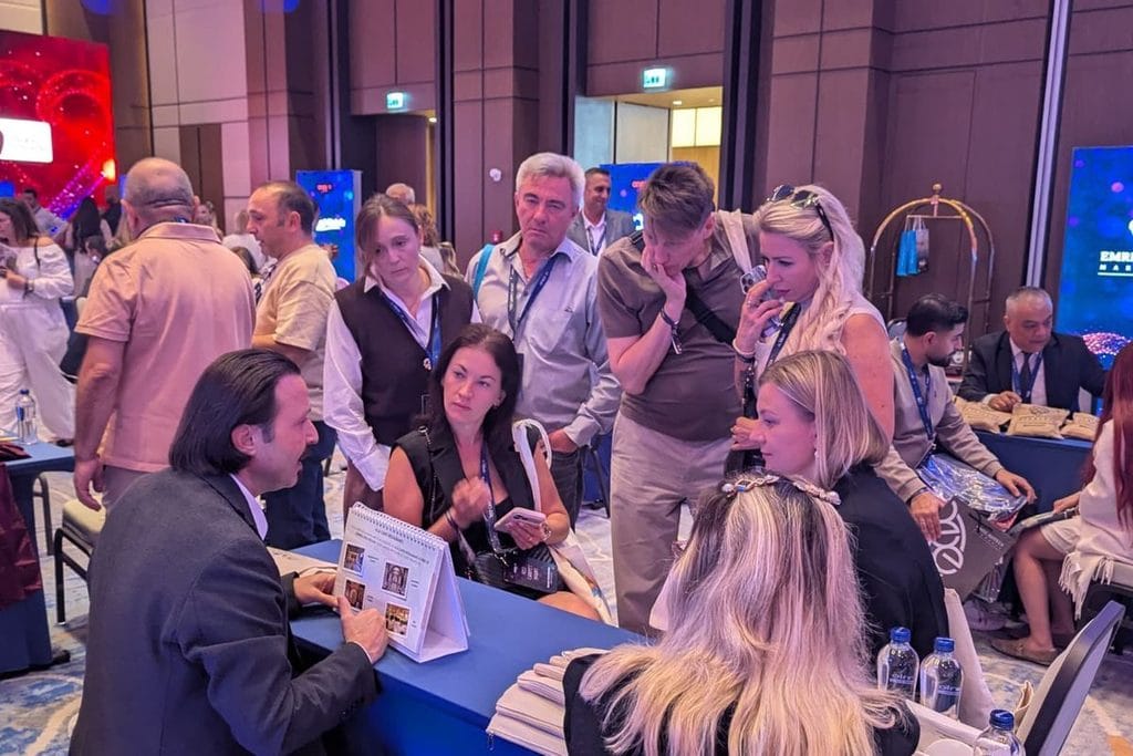 Eine Gruppe von Menschen steht in einem Konferenzraum um einen blauen Tisch herum, während ein Mann an einem Tisch eine Broschüre präsentiert und erklärt.
