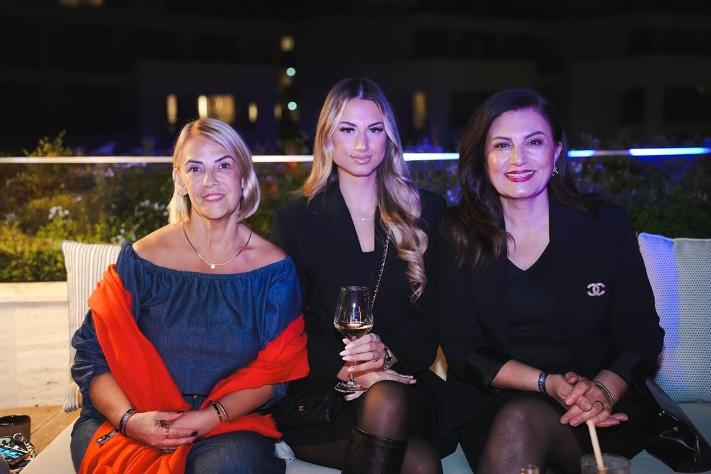 Drei Frauen sitzen abends auf einer Terrasse nebeneinander. Die linke trägt ein blaues Oberteil mit rotem Tuch, die anderen beiden sind in Schwarz gekleidet.