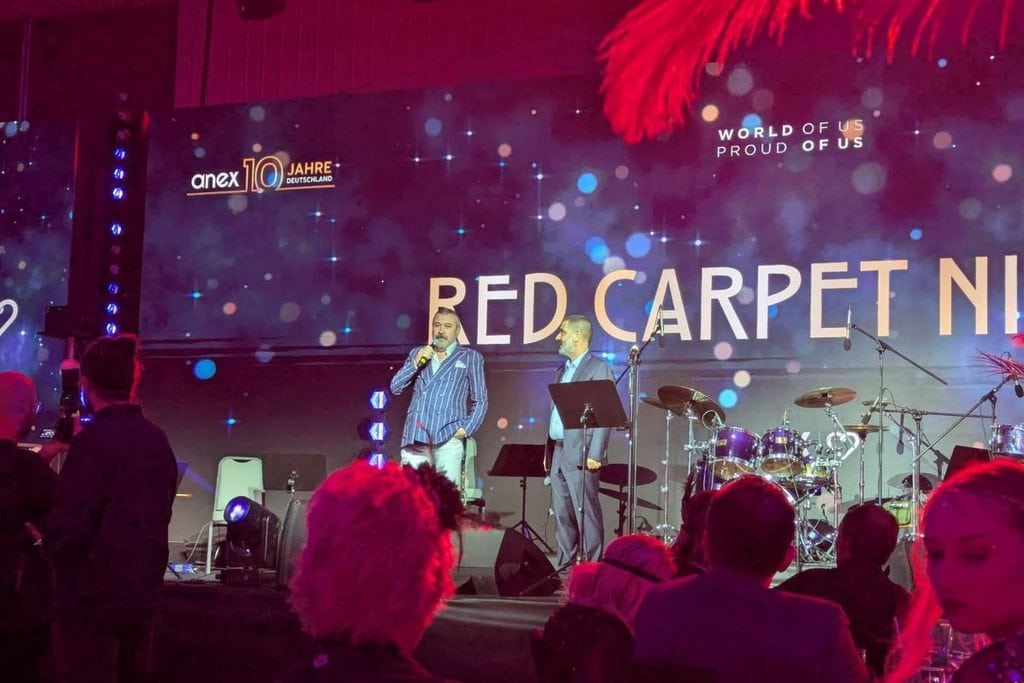 Zwei Männer stehen auf einer Bühne vor einer großen Leinwand mit der Aufschrift Red Carpet Night, während ein Publikum die Szene von unten betrachtet.
