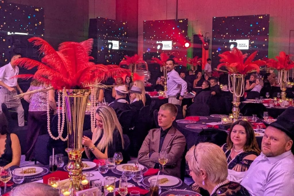 Festlich gedeckte Tische in einem Saal mit großen roten Federgestecken in Goldvasen und Bildschirmen im Hintergrund, an denen Gäste bei einem Event sitzen.