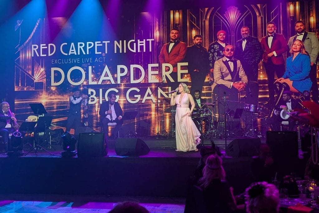 Eine Sängerin in einem langen hellen Kleid steht auf einer Bühne vor einer großen Videowand, die eine Band zeigt, während Musiker sie live begleiten.