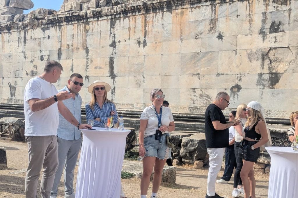 Eine Gruppe von Menschen steht vor einer antiken Steinmauer an Stehtischen mit weißen Hussen, einige Personen tragen Sonnenbrillen und legere Kleidung.