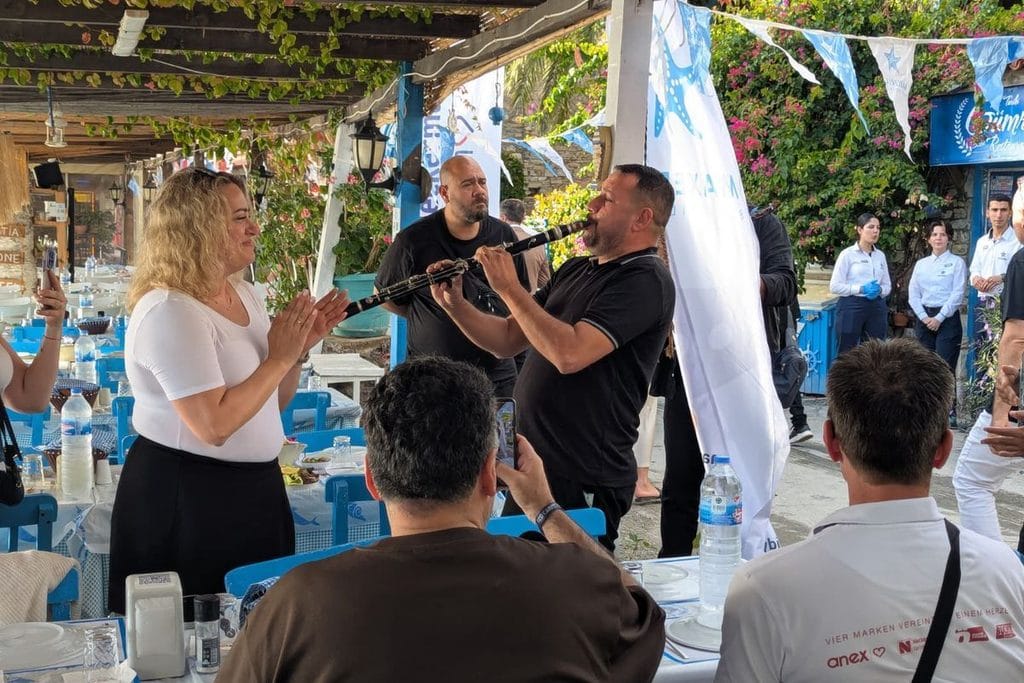 Eine Frau klatscht im Takt, während ein Mann in einem Restaurant im Freien Klarinette spielt. Gäste sitzen an gedeckten Tischen und beobachten die Szene.