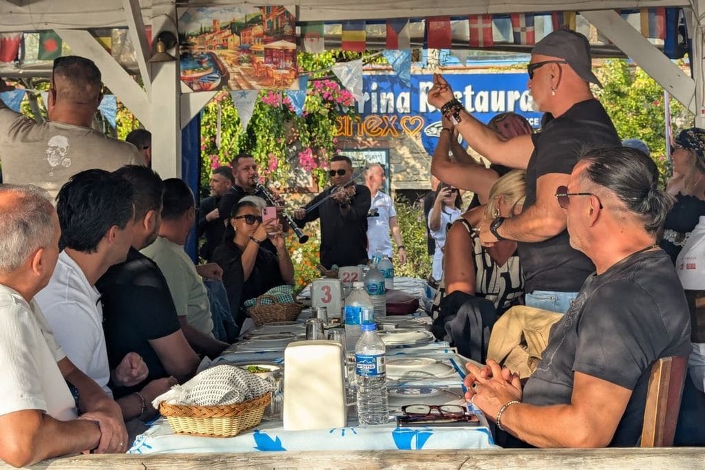 Eine Gruppe von Menschen sitzt an einem gedeckten Tisch in einem Restaurant im Freien, während im Hintergrund zwei Musiker Geige und Klarinette spielen.