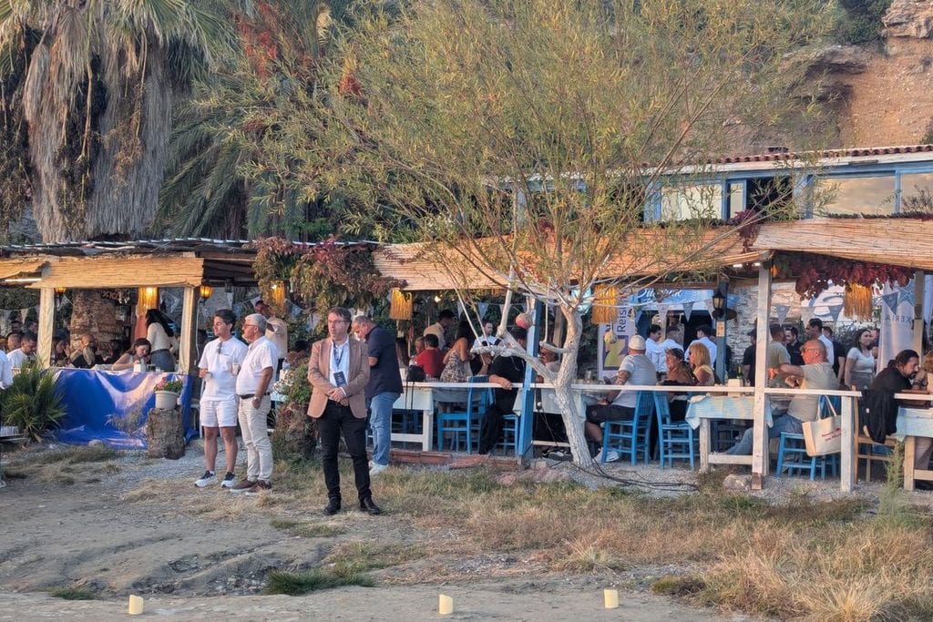 Eine Gruppe von Menschen steht und sitzt in einem Restaurant im Freien mit Strohdächern, umgeben von Bäumen und Felsen in der Abenddämmerung im Freien.