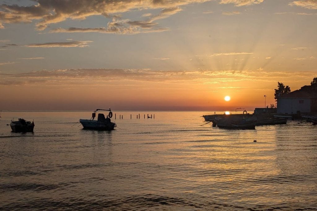 Mehrere kleine Fischerboote liegen bei Sonnenuntergang in einer ruhigen Bucht vor Anker, während das goldene Licht den Horizont und das Wasser beleuchtet.