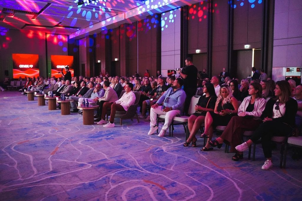 Ein großes Publikum sitzt in einem festlich beleuchteten Konferenzsaal mit gemustertem Teppichboden und blickt auf eine Bühne mit großen digitalen Displays.