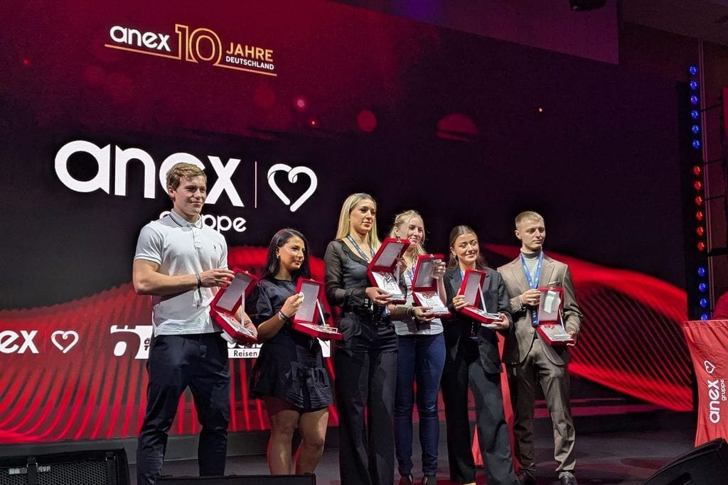 Sechs Personen stehen auf einer Bühne vor einer großen Leinwand mit dem Logo der anex Gruppe und halten geöffnete Auszeichnungsboxen in ihren Händen.