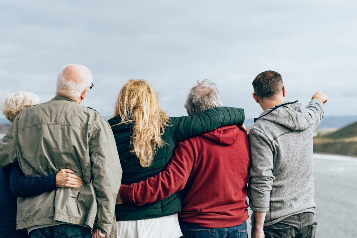 Eine Gruppe von vier Personen steht mit dem Rücken zur Kamera und blickt auf eine Küstenlandschaft, wobei eine Person in die Ferne auf das Meer zeigt.