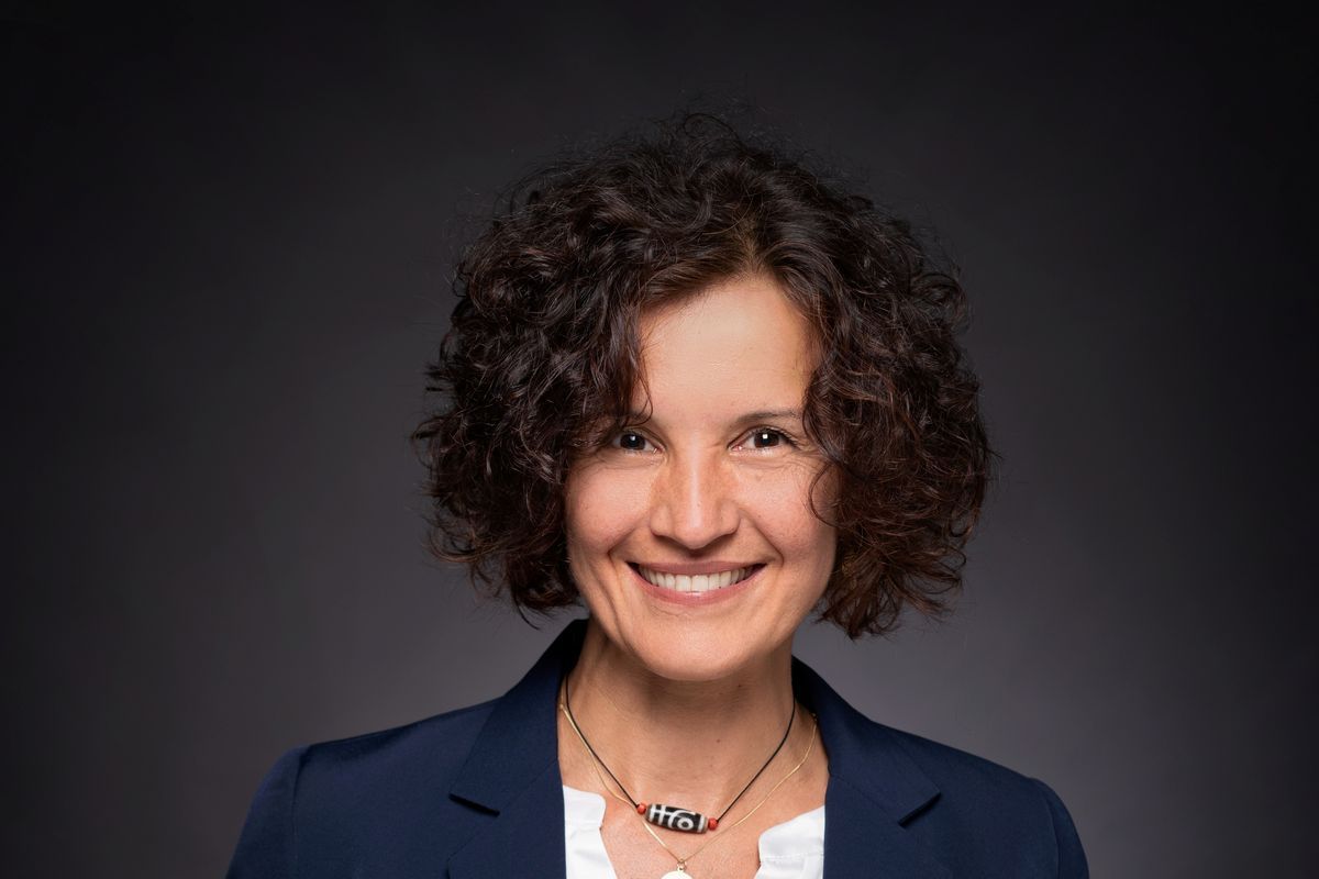 Eine lächelnde Frau mit dunklen, lockigen Haaren trägt ein weißes Oberteil unter einem dunkelblauen Blazer vor einem neutralen, grauen Hintergrund.