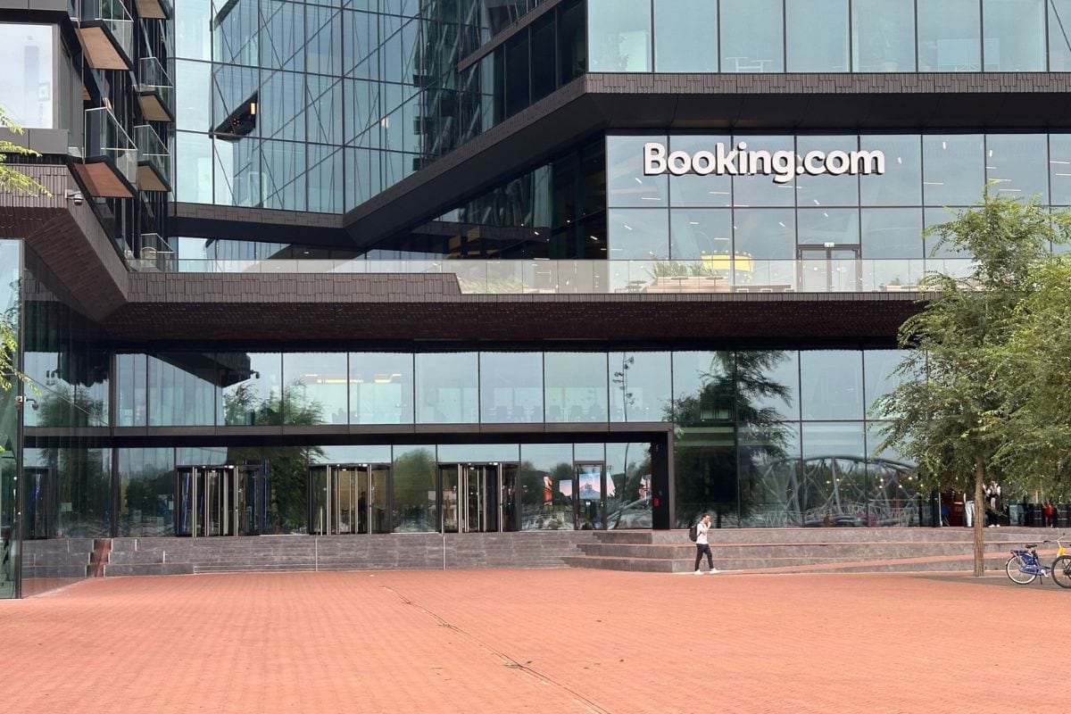 Die moderne Glasfassade des Booking.com-Gebäudes mit dem Firmenlogo, davor ein gepflasterter Vorplatz, eine Person und ein Fahrrad bei Tageslicht im Freien.