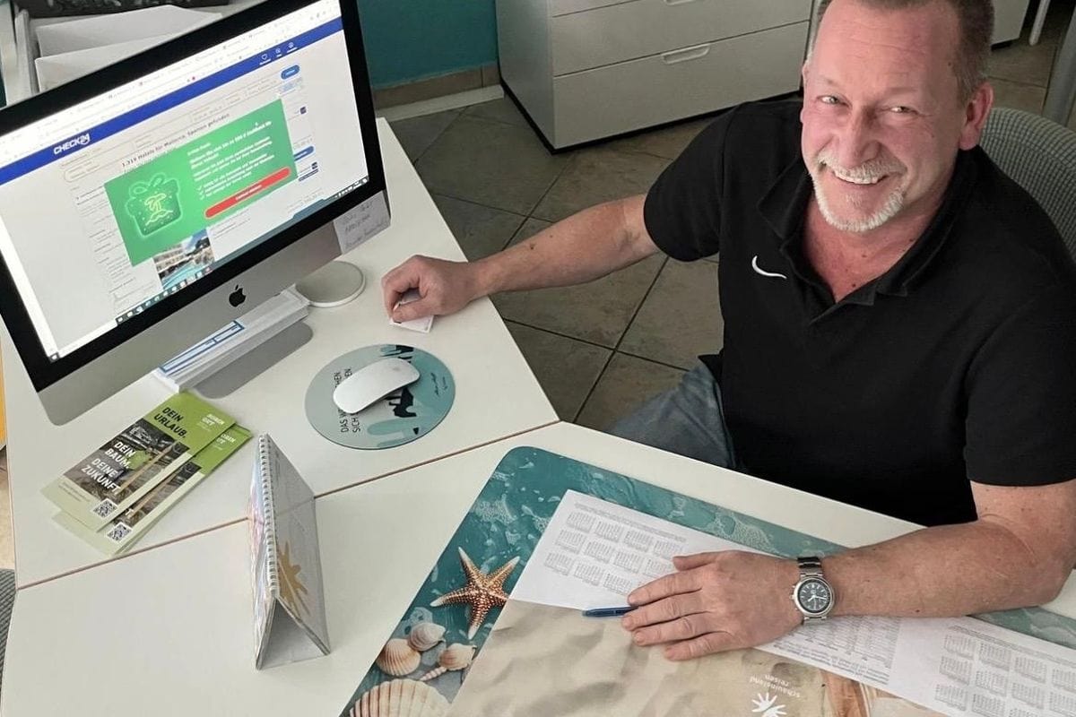 Ein lächelnder Mann sitzt an einem Schreibtisch vor einem Computerbildschirm, auf dem die Website von CHECK24 mit Suchergebnissen für Mallorca zu sehen ist.